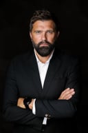 Bearded man with arms crossed in a black suit against a dark studio background