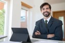Smiling bearded man in a pinstripe suit sitting at his desk with a tablet