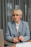 Short-haired blonde businesswoman in a grey-blue blazer seated at her desk